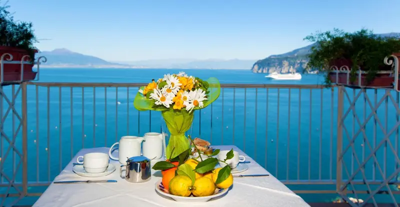 Une table est dressée avec un service à thé et des fruits frais sur une terrasse ensoleillée.