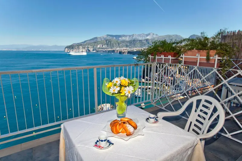 Une table avec un petit-déjeuner est mise en scène sur une terrasse offrant une vue sur la mer et les montagnes.