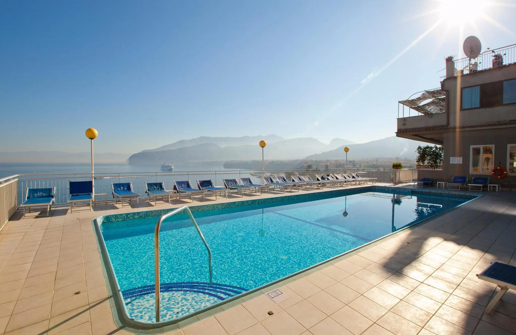 Une piscine bleue bordée de transats ensoleillés offre une vue sur la mer et les montagnes.