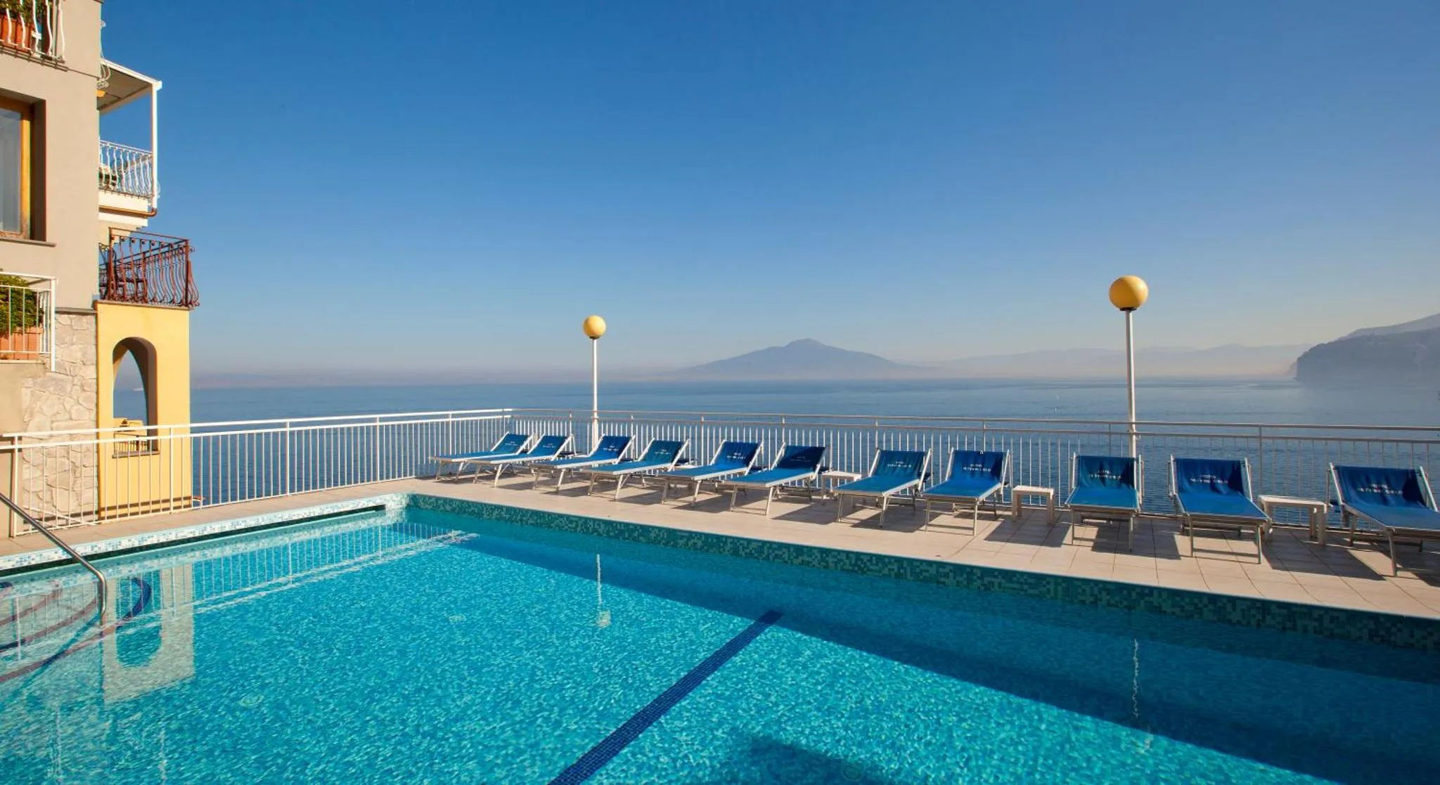 Transats bleus alignés au bord d'une piscine turquoise avec vue sur la mer.