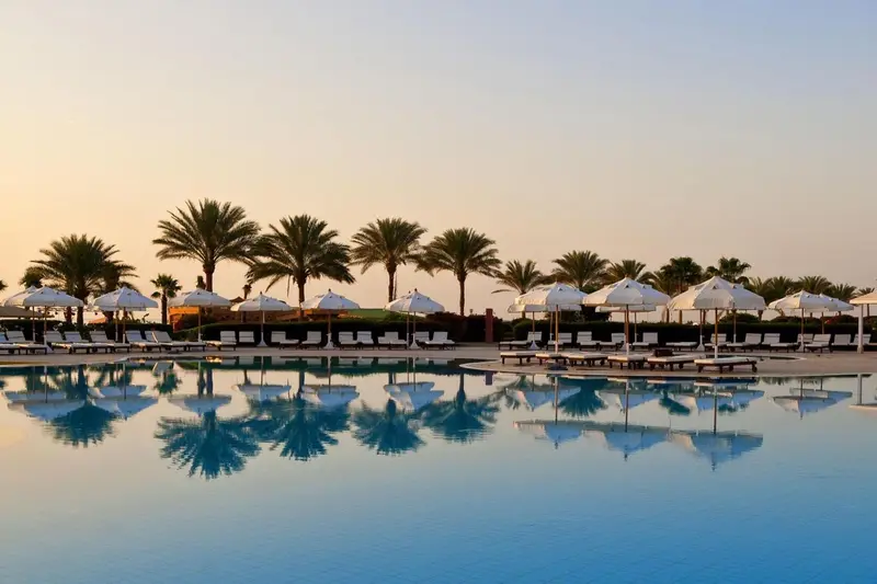 Transats et parasols bordent une piscine turquoise reflétant le ciel et la végétation.