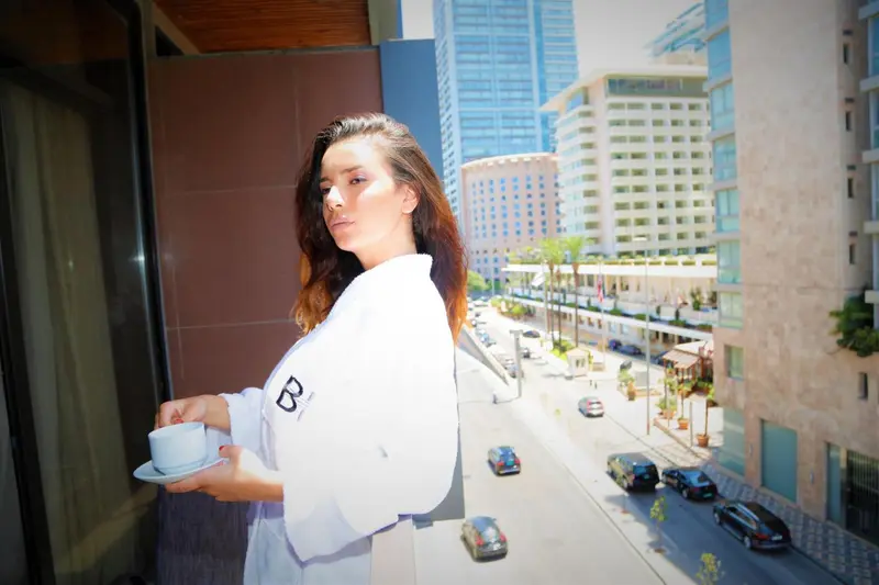 Une femme en peignoir tient une tasse sur un balcon avec vue sur la ville.