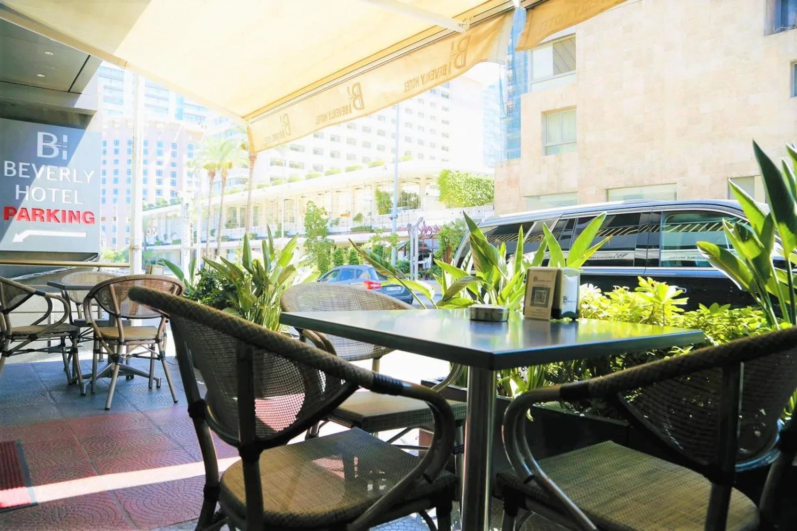 Tables et chaises disposées sur une terrasse ombragée devant un bâtiment.
