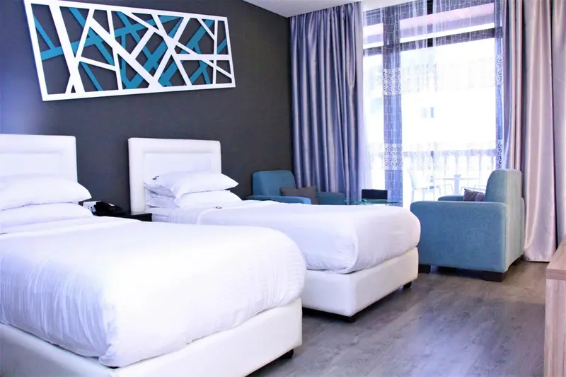 Chambre lumineuse avec deux lits simples et un fauteuil turquoise.