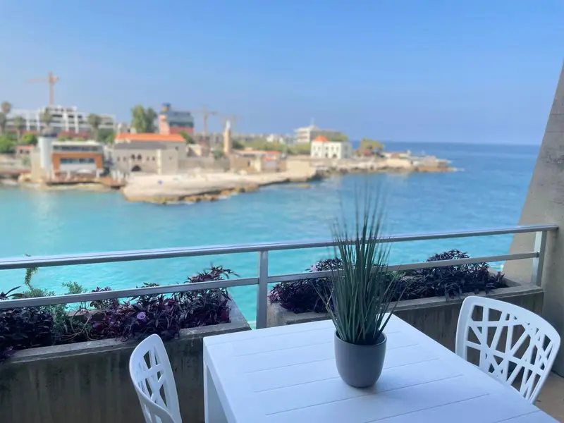 Une table et des chaises blanches sur un balcon avec vue sur la mer et la côte rocheuse.