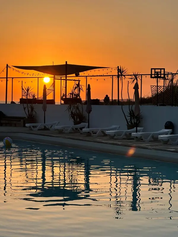 Une piscine reflète le ciel orangé au coucher du soleil, avec des transats et des palmiers en arrière-plan.