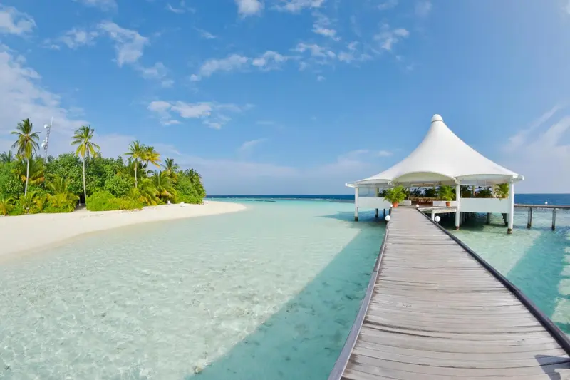 Un ponton en bois mène à un pavillon blanc sur une eau turquoise et une plage de sable clair.