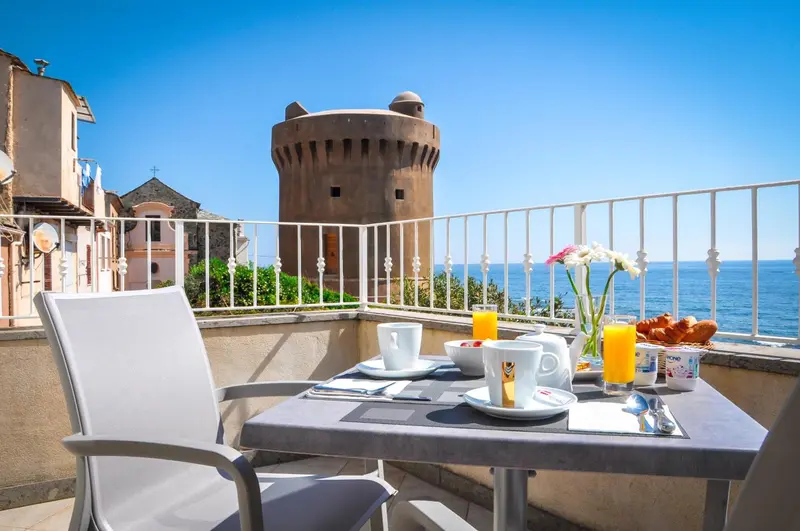 Une table de petit-déjeuner est dressée sur une terrasse avec vue sur la mer et un monument historique.