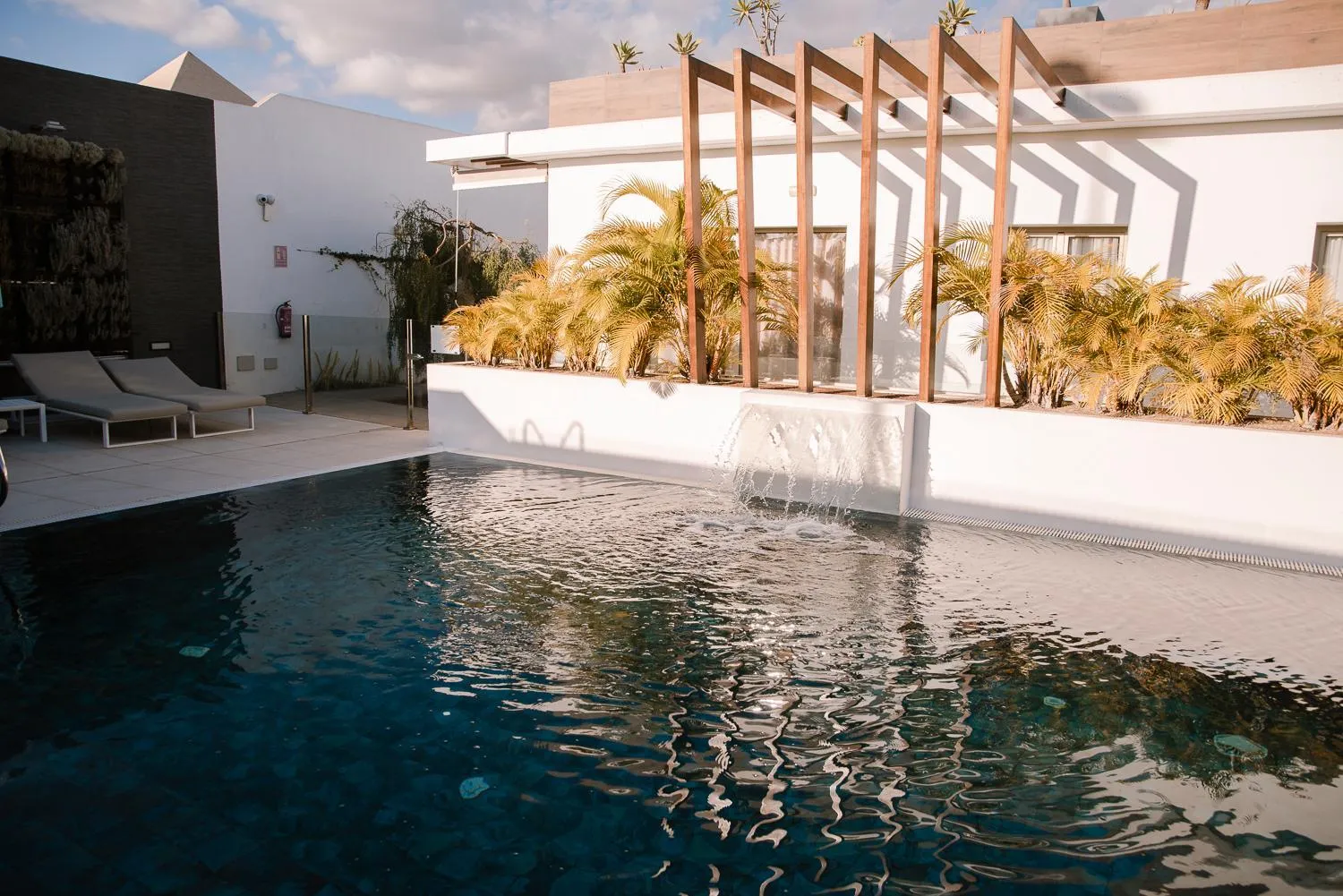 Une piscine rectangulaire avec une fontaine et des transats sur une terrasse ensoleillée.