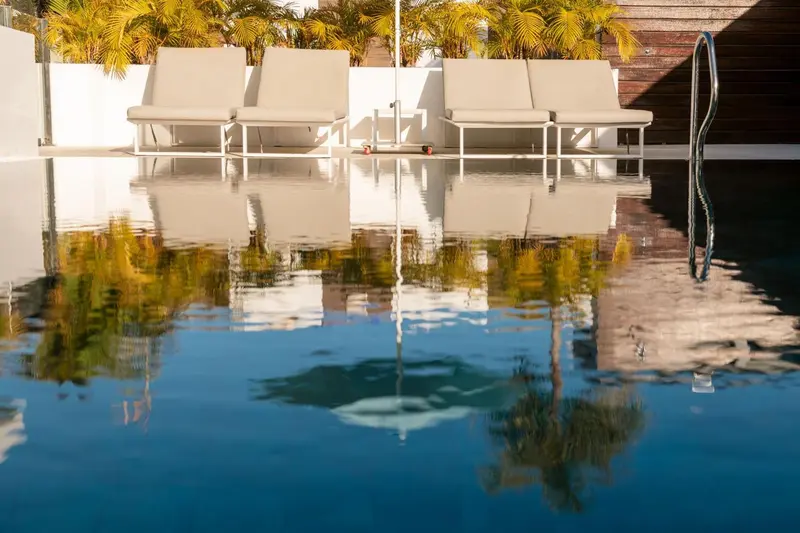 Transats blancs alignés au bord d'une piscine turquoise reflétant la végétation.