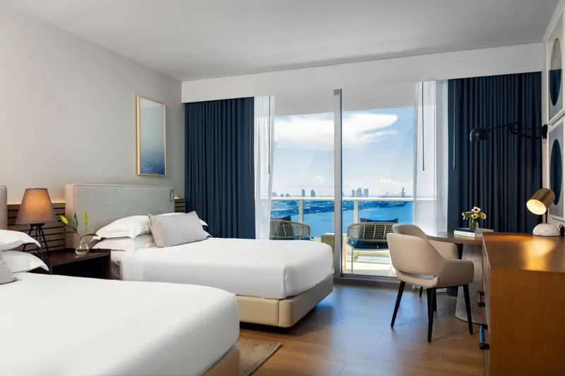 Chambre lumineuse avec deux lits et vue sur la mer depuis un balcon.