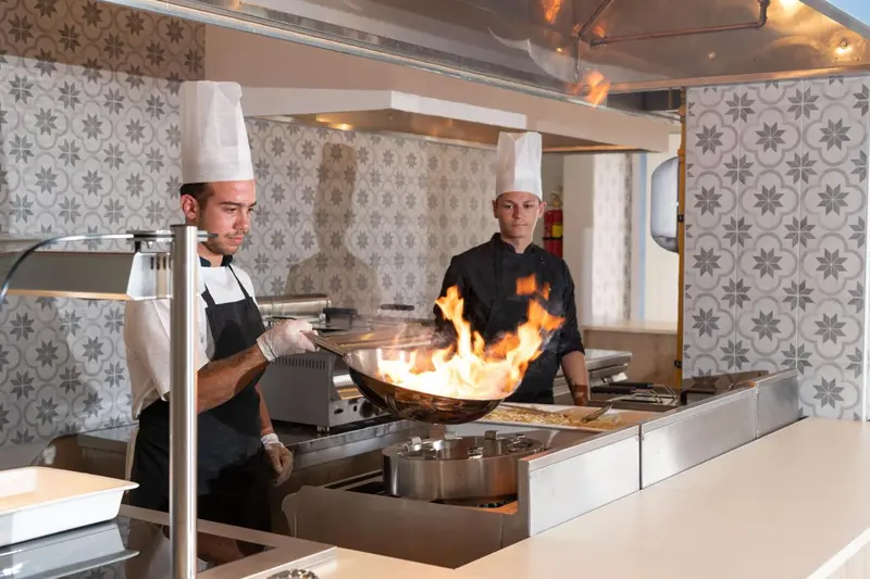 Deux cuisiniers préparent un plat avec des flammes dans une cuisine professionnelle.
