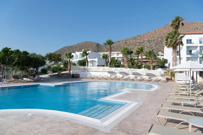Piscine rectangulaire entourée de transats et de palmiers, avec un bâtiment blanc en arrière-plan.