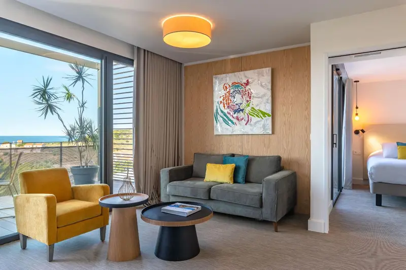 Un salon lumineux avec canapé et fauteuil face à une baie vitrée donnant sur la mer.