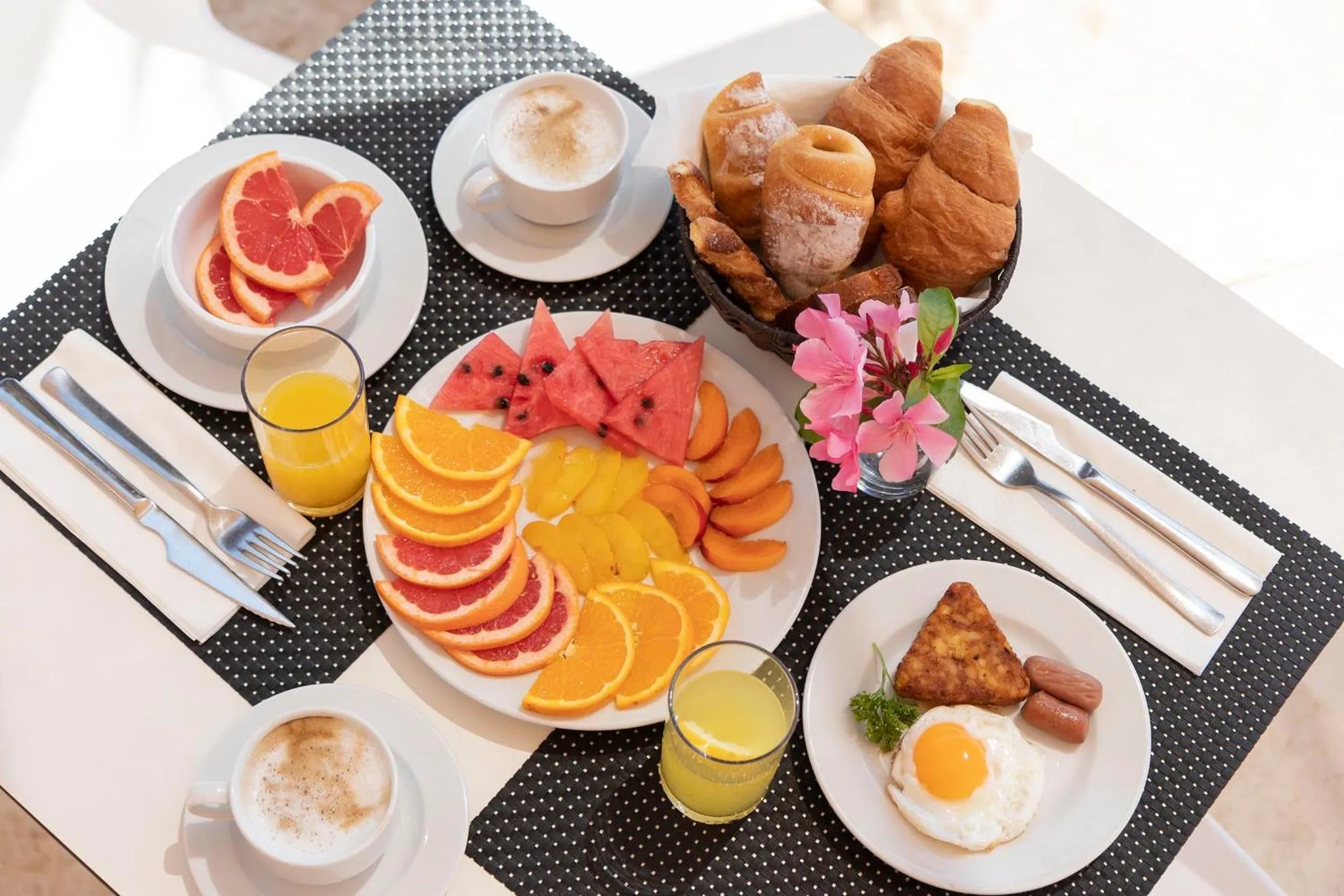Une table blanche est dressée avec un copieux petit-déjeuner composé de fruits frais, de viennoiseries et de boissons.