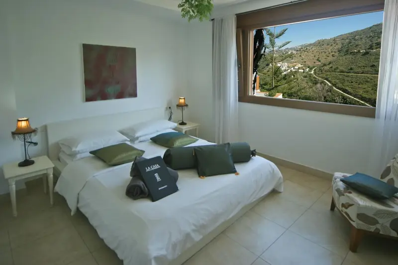 Une chambre lumineuse avec un grand lit recouvert de coussins, donnant sur un paysage montagneux.