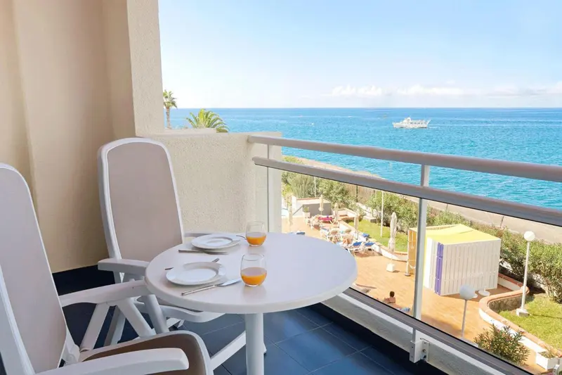 Une table ronde avec des couverts et des boissons sur un balcon ensoleillé avec vue sur la mer.