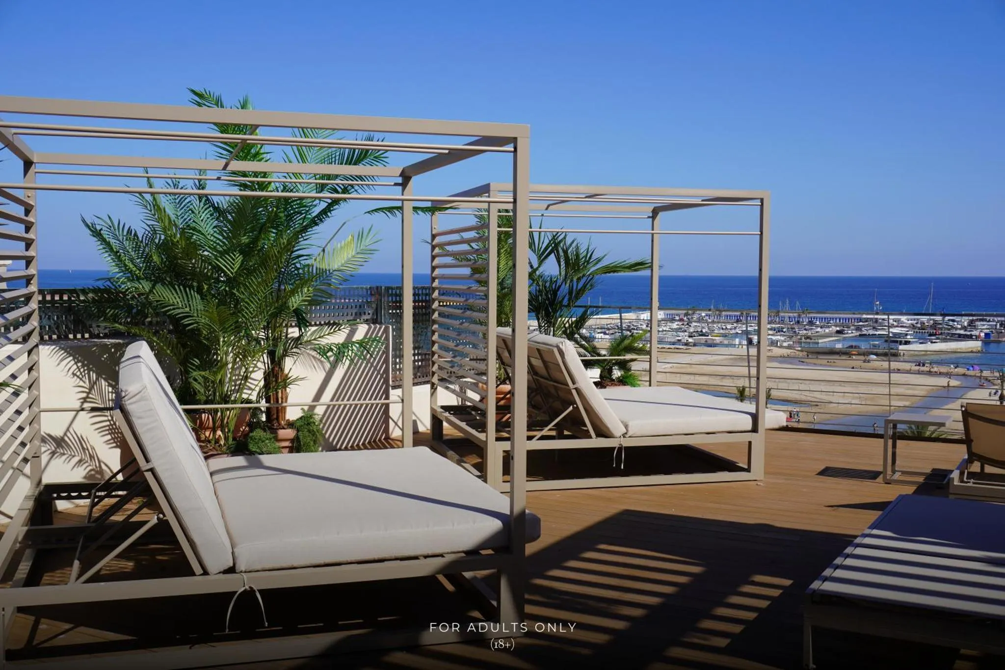 Transats et pergolas sur une terrasse ensoleillée avec vue sur la mer et un port de plaisance.