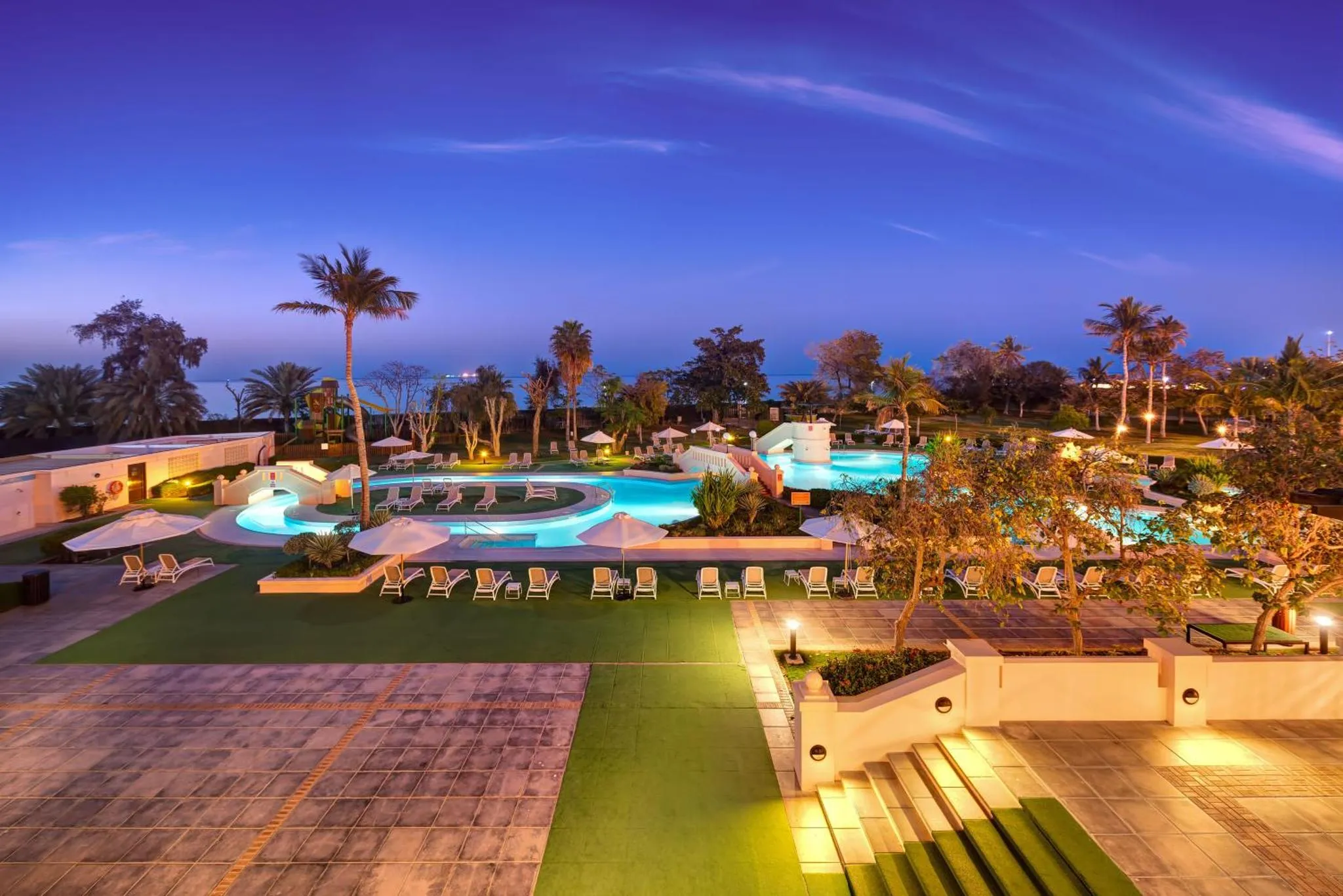Vue d'une piscine illuminée au crépuscule, entourée de palmiers et de transats.