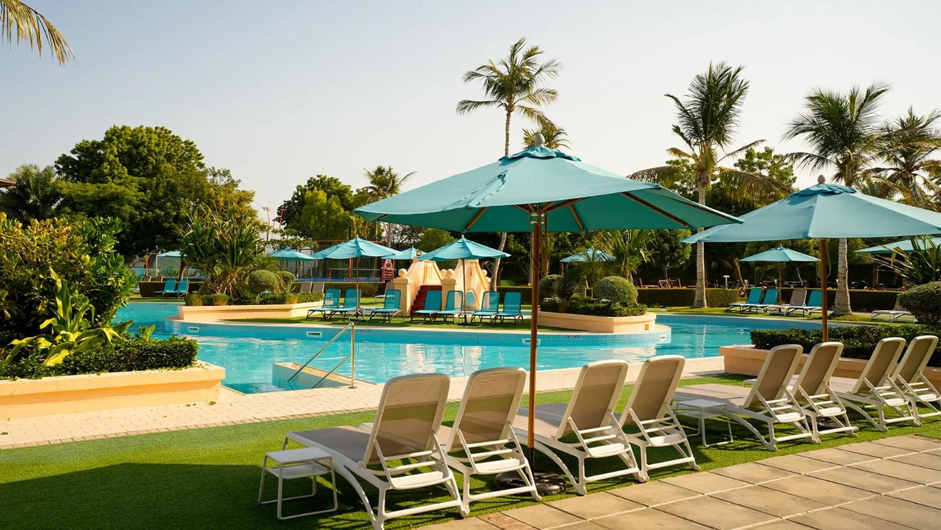Transats blancs disposés au bord d'une piscine turquoise ombragée par des parasols.