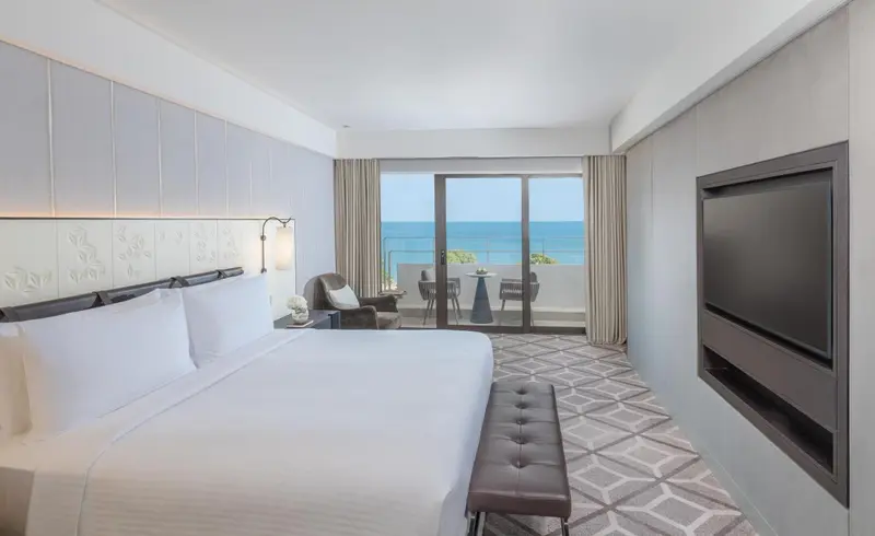 Chambre lumineuse avec un grand lit et une vue sur la mer depuis un balcon.