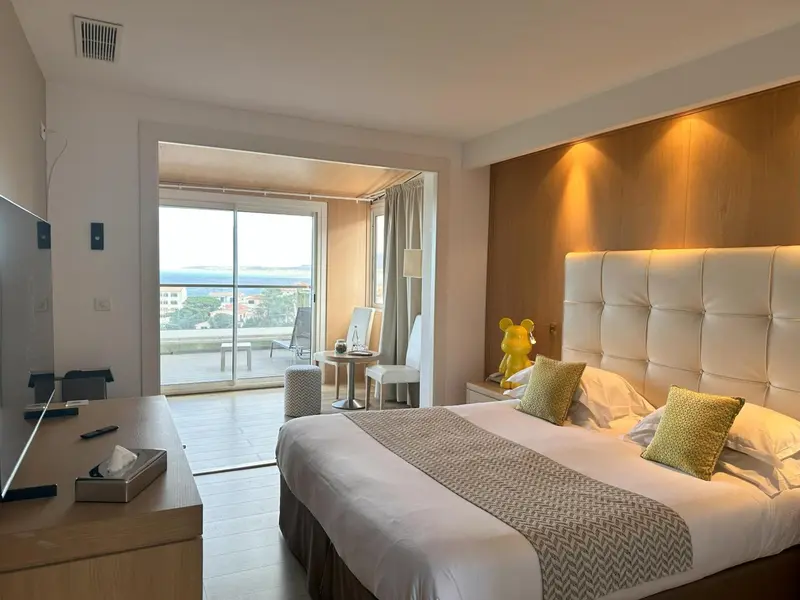 Chambre lumineuse avec un grand lit et une vue sur la mer depuis une terrasse.