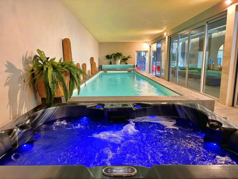 Un jacuzzi bleu illuminé au premier plan, avec une piscine intérieure et des plantes vertes en arrière-plan.
