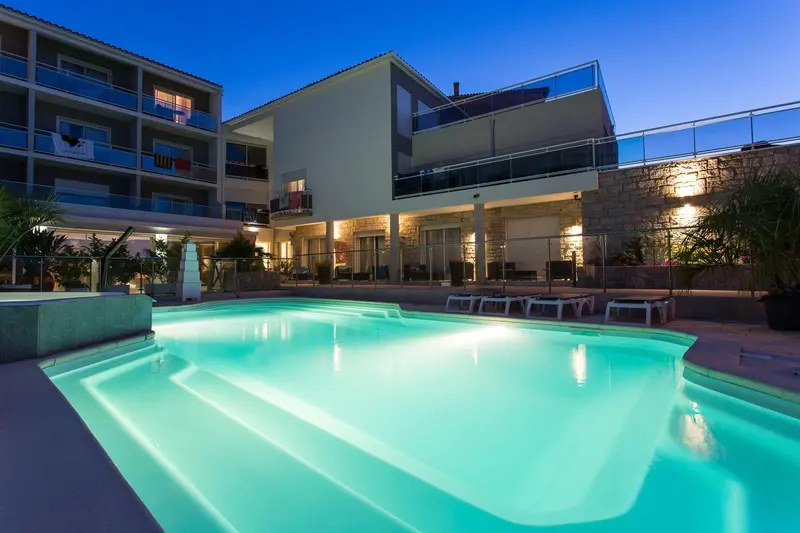 Piscine illuminée devant un bâtiment moderne avec des balcons.
