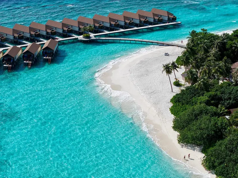 Vue aérienne d'une plage de sable blanc bordée d'eau turquoise et de bungalows sur pilotis.