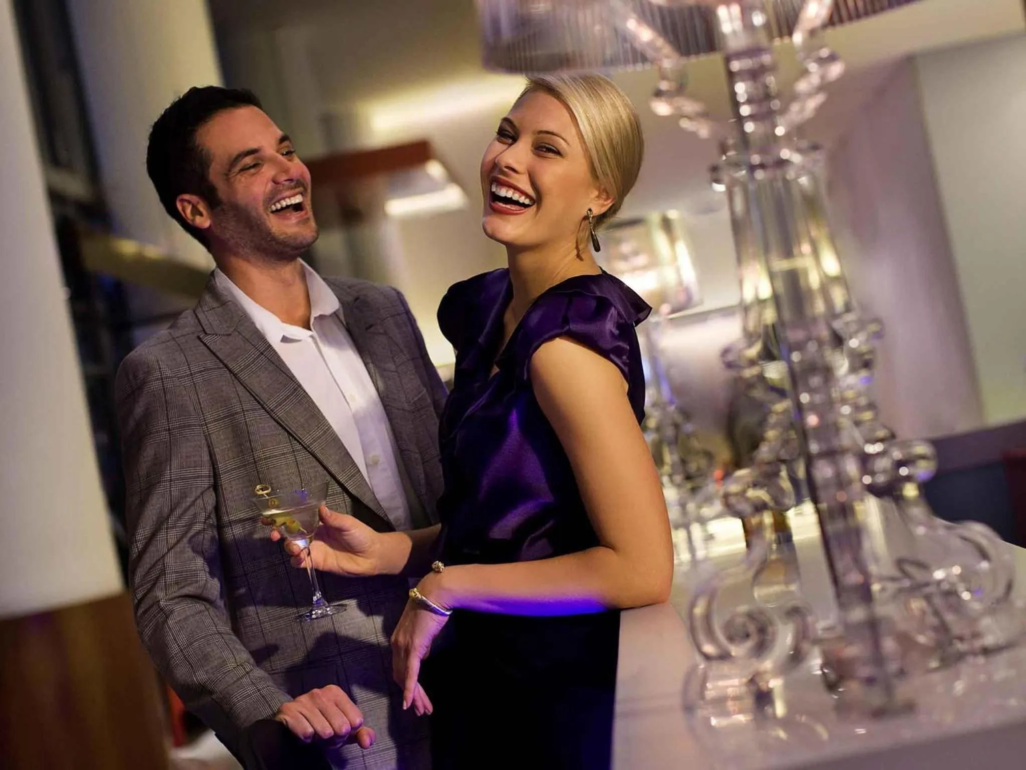 Un couple rit en tenant des verres à cocktail dans un espace lumineux.