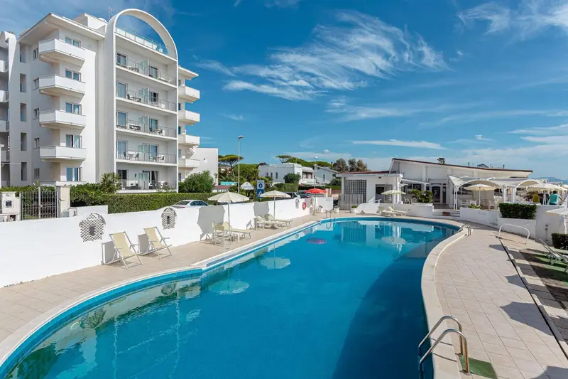 Piscine rectangulaire turquoise devant un bâtiment blanc et une végétation luxuriante sous un ciel bleu.