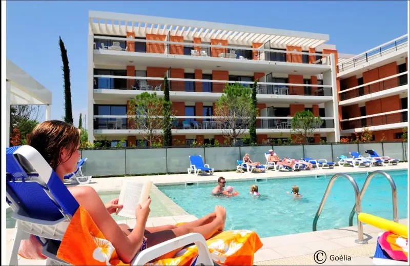 Une personne se relaxe au bord d'une piscine ensoleillée devant un bâtiment blanc.