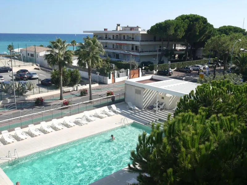 Vue aérienne d'une piscine d'hôtel bordée de transats, avec la mer en arrière-plan.
