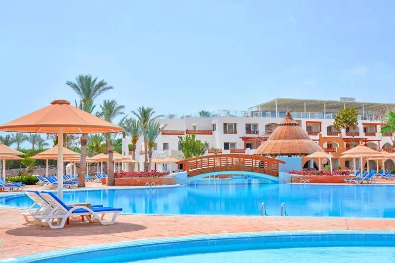 Transats bleus et parasols orangés bordent une piscine turquoise devant un bâtiment blanc.