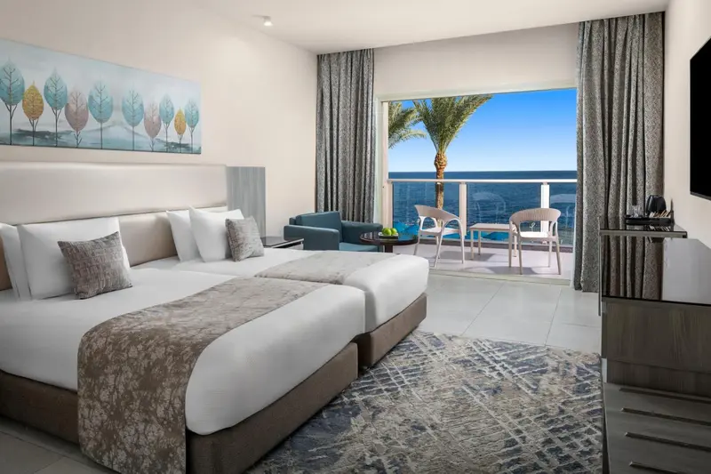 Chambre lumineuse avec un grand lit et un accès à un balcon offrant une vue sur la mer.