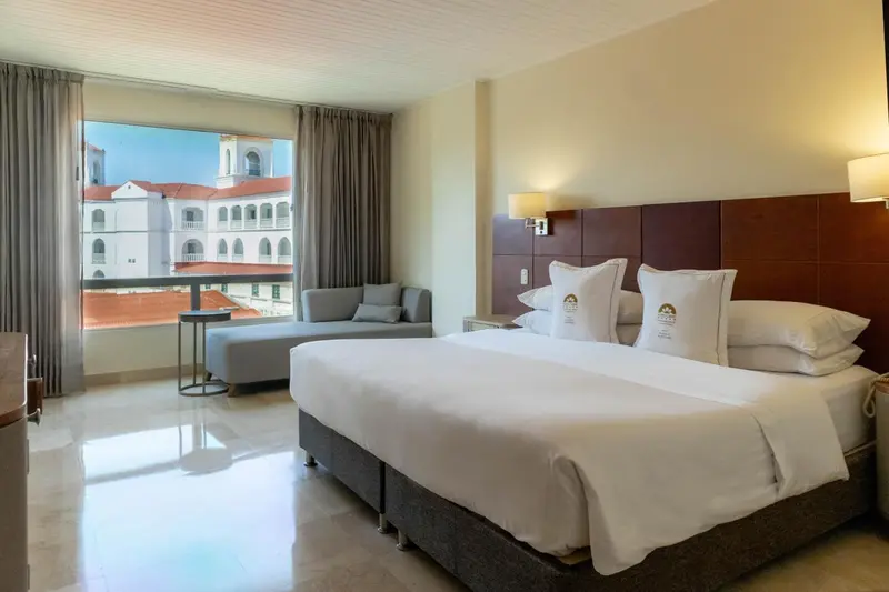 Chambre lumineuse avec un grand lit et un coin salon près d'une fenêtre.