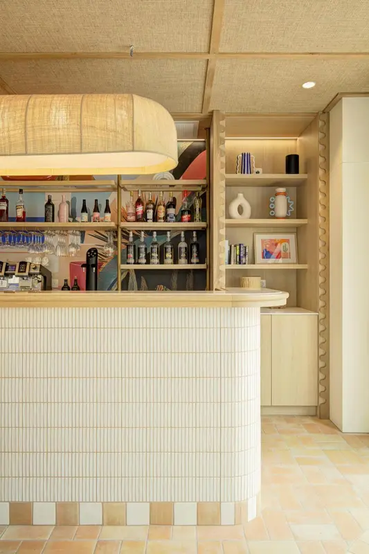 Un comptoir de bar en mosaïque blanche est éclairé par une suspension en fibres naturelles.