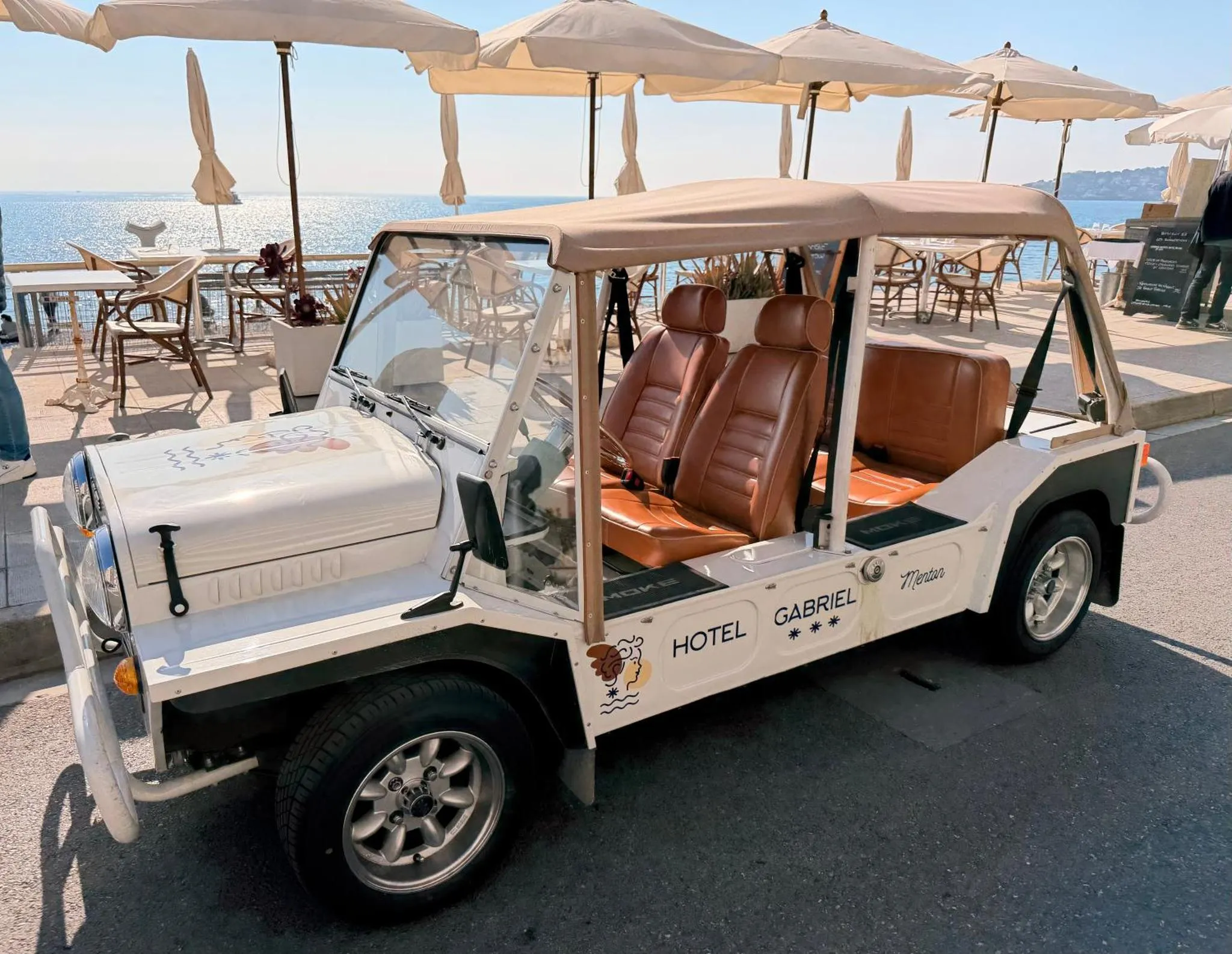 Un véhicule blanc ouvert se trouve sur un parking ensoleillé face à la mer.