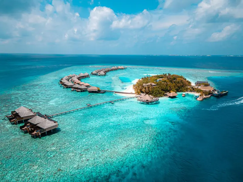 Vue aérienne d'un îlot tropical avec des bungalows sur pilotis et une eau turquoise.