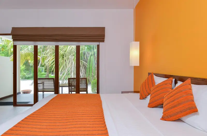 Une chambre lumineuse avec un lit recouvert d'une courtepointe orange et de coussins assortis, donnant sur une terrasse et une végétation tropicale.