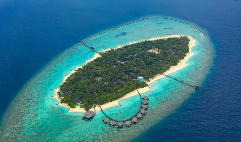 Vue aérienne d'une île tropicale entourée d'un lagon turquoise et de bungalows sur pilotis.