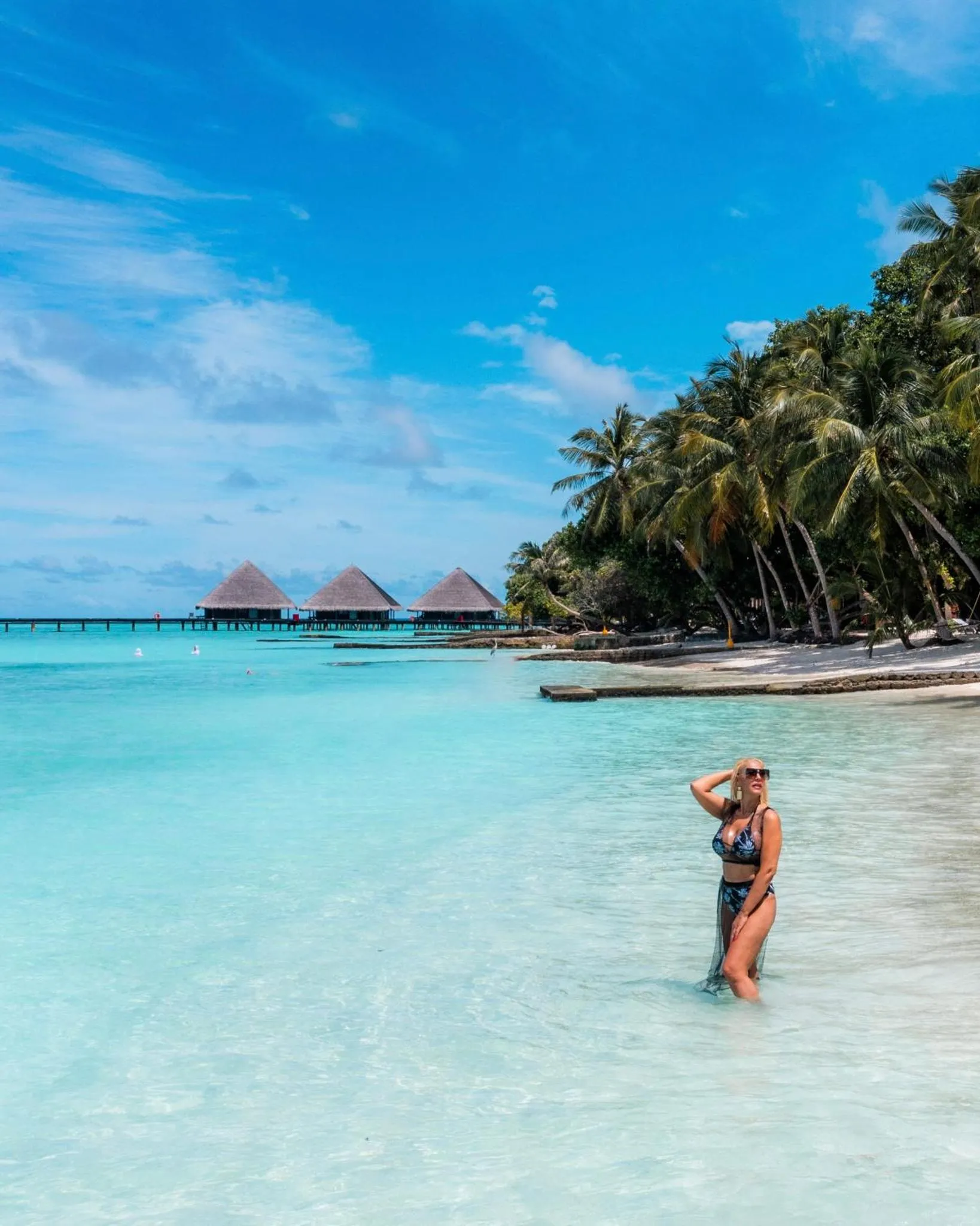 Une femme se tient dans l'eau turquoise devant une plage de sable blanc et des bungalows sur pilotis.