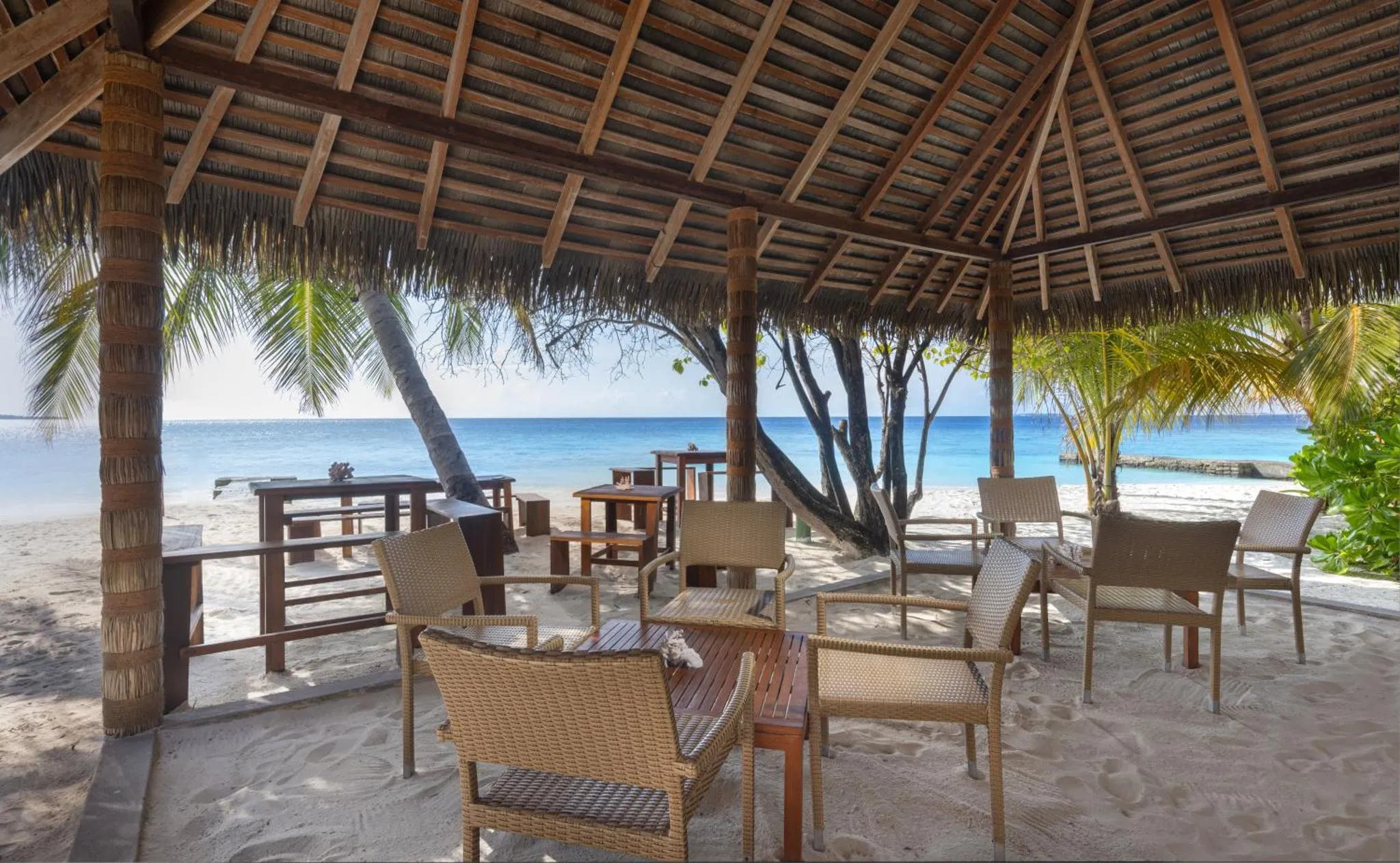 Espace de détente ombragé avec mobilier en rotin face à une plage de sable blanc.