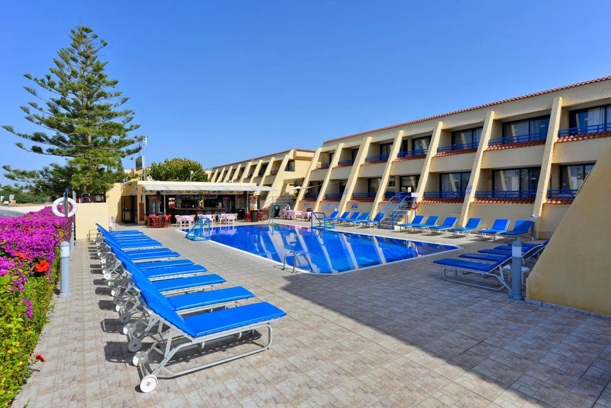 Transats bleus alignés au bord d'une piscine ensoleillée devant un bâtiment jaune.