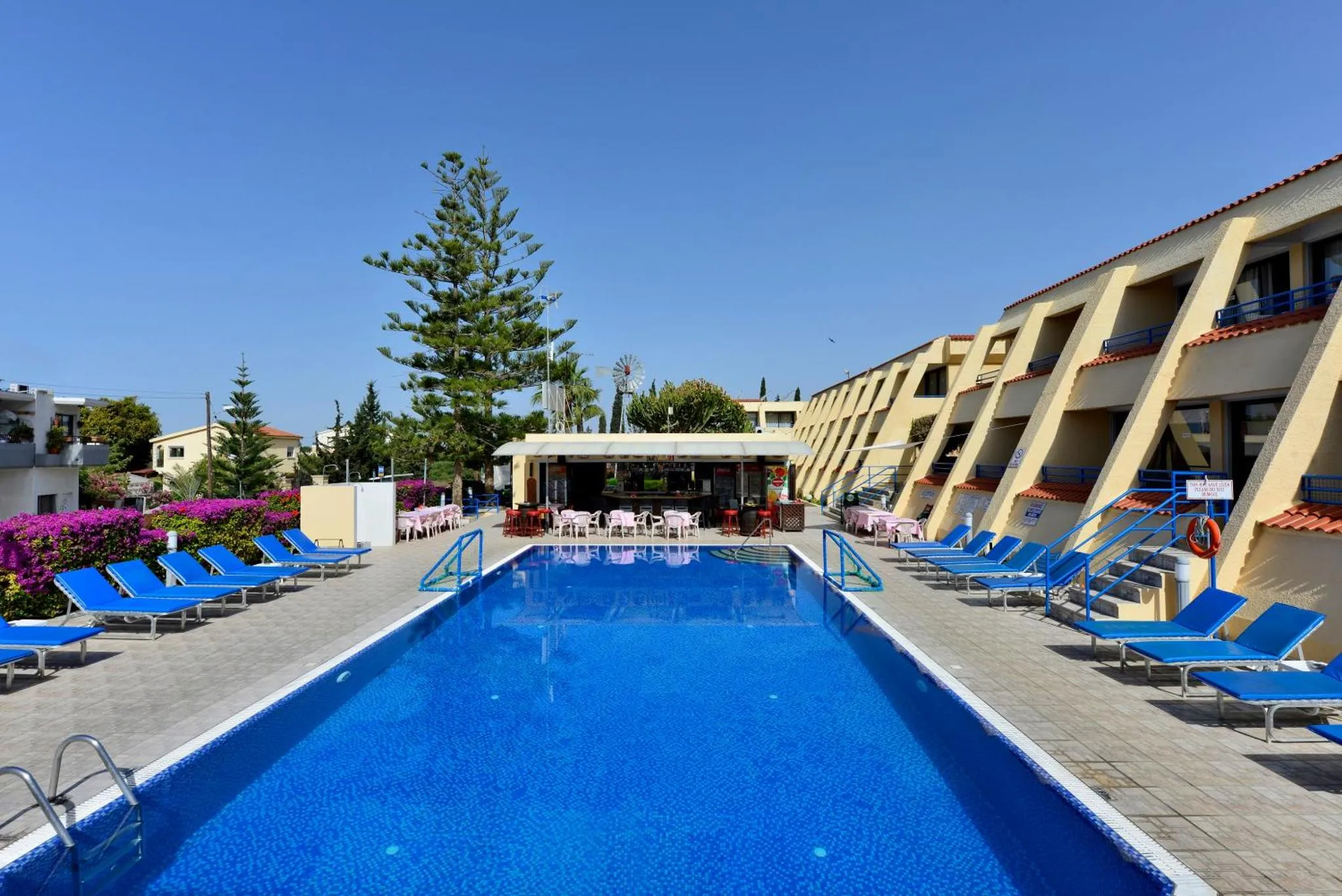 Piscine rectangulaire bordée de transats bleus devant un bâtiment à deux étages.