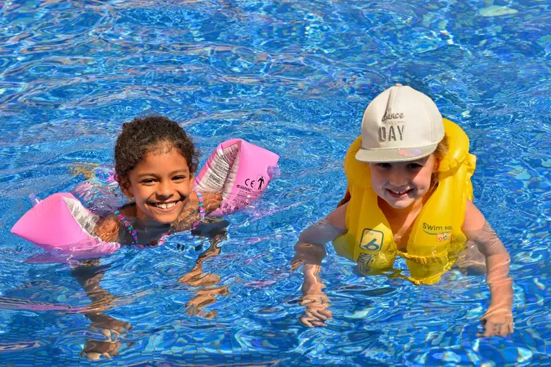 Deux enfants sourient dans une piscine ensoleillée, portant des dispositifs de flottation.