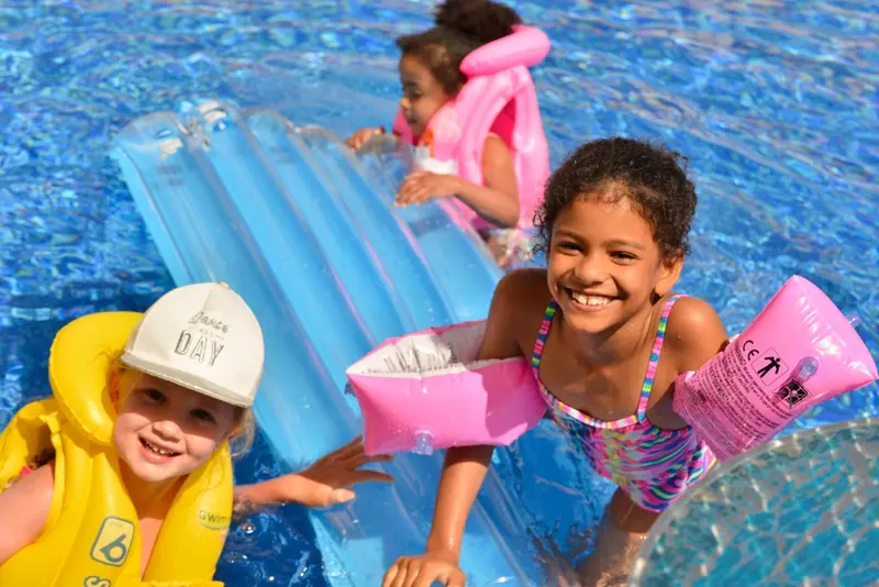 Des enfants sourient et s'amusent dans une piscine avec des bouées et un toboggan.