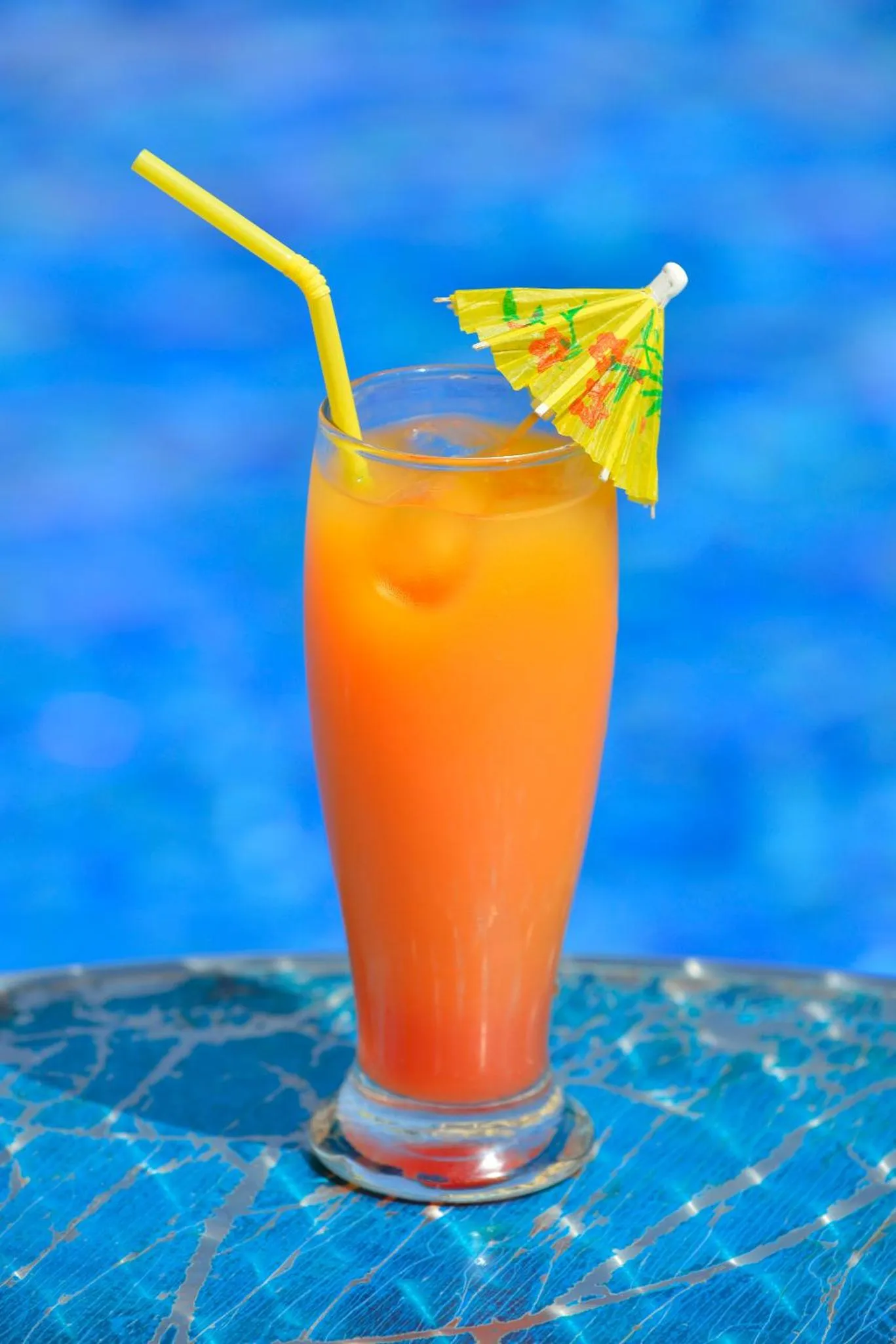 Un cocktail orangé dans un verre décoré, posé sur une table près d'une piscine.