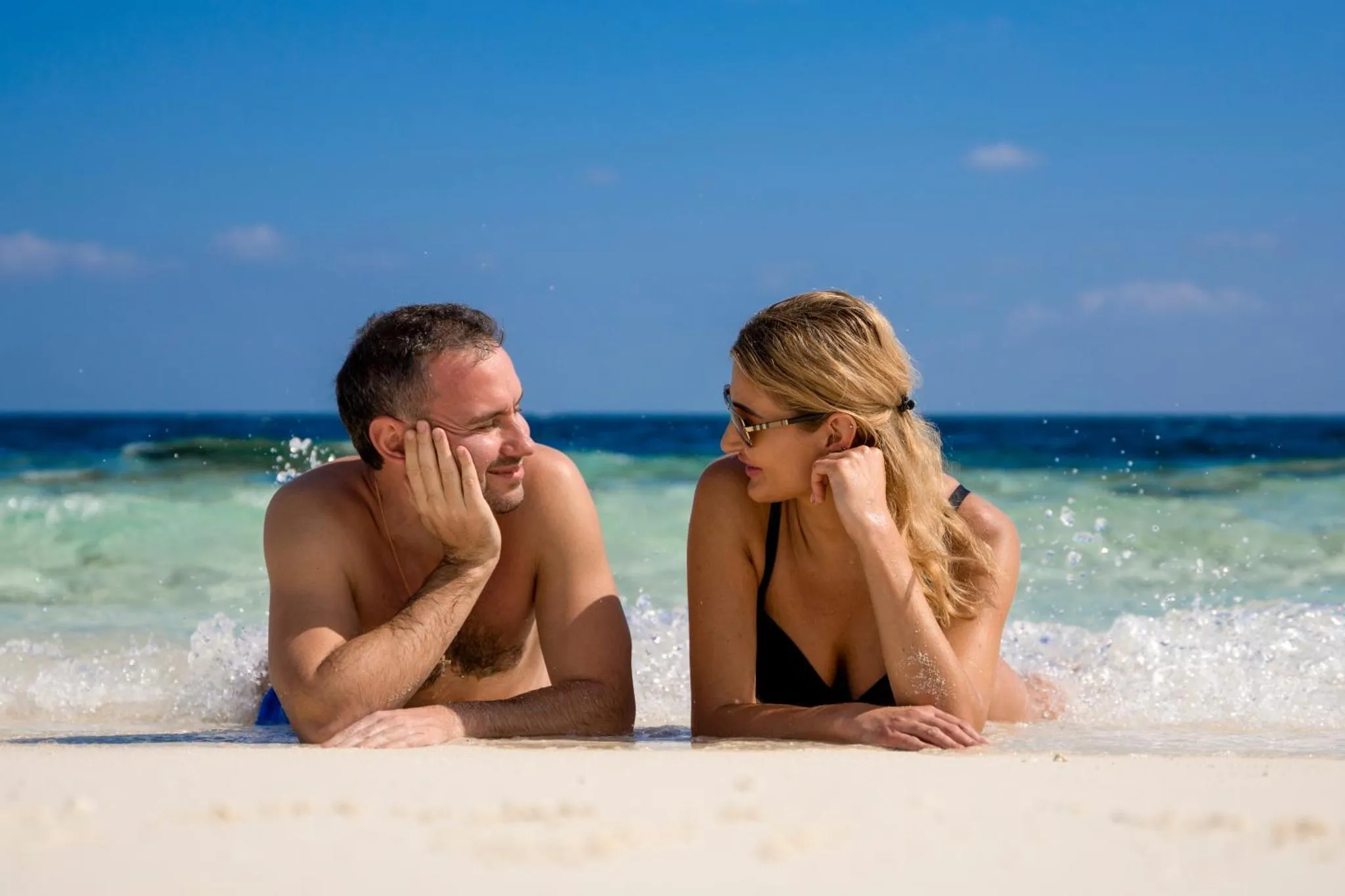 Un couple souriant se détend allongé dans les vagues sur une plage de sable blanc.