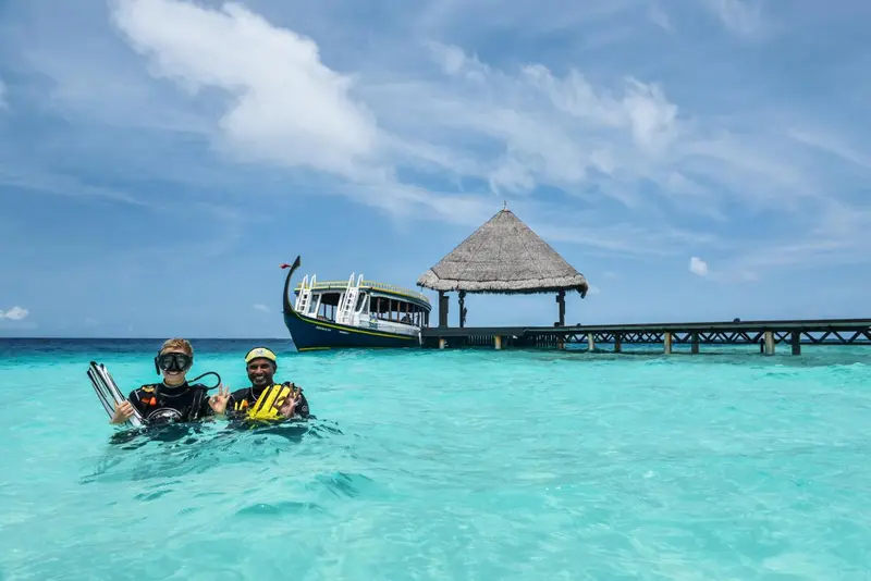Deux personnes sourient dans une eau turquoise, près d'une structure sur pilotis.